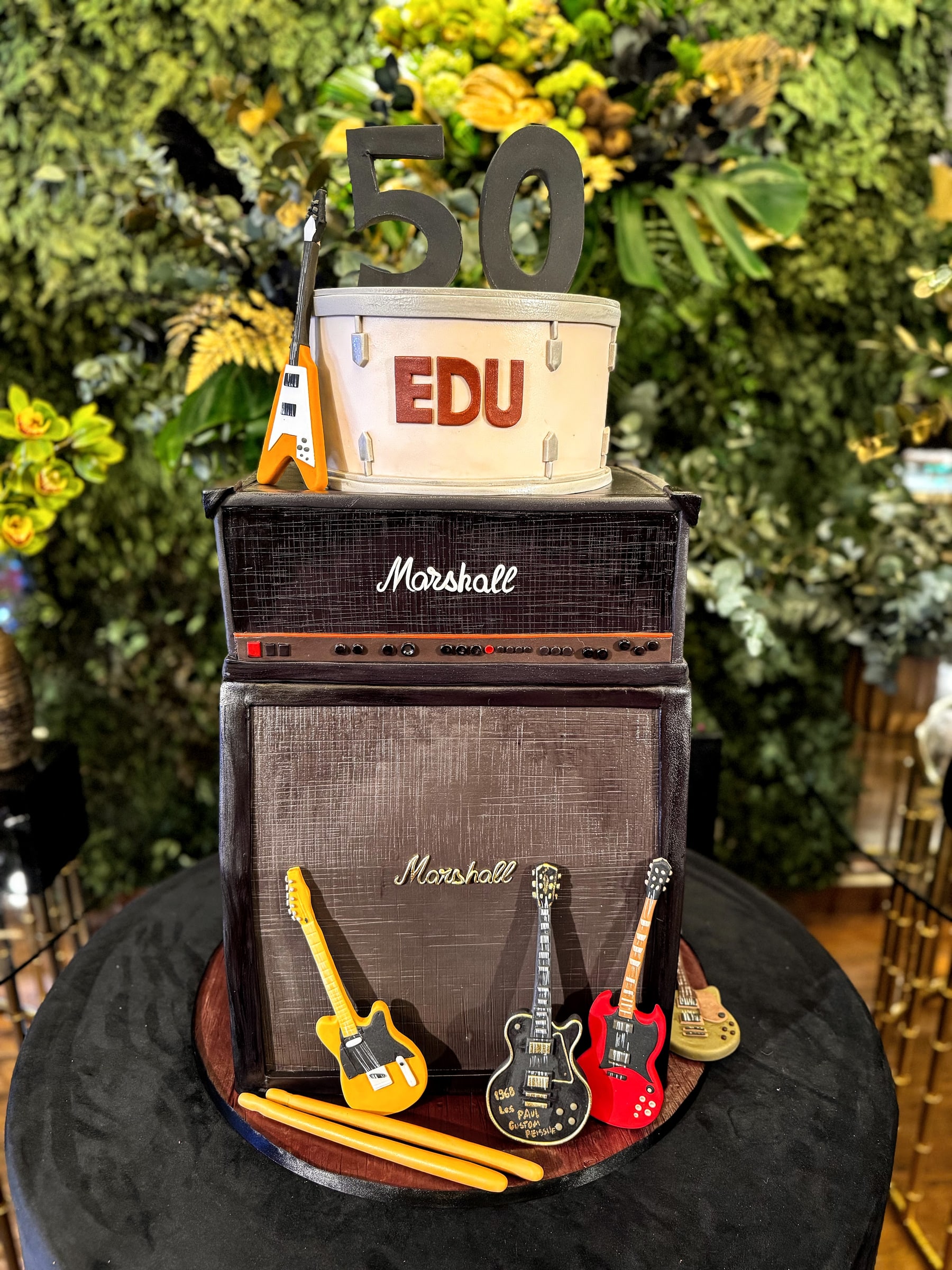 Bolo escultórico de aniversário de 50 anos em formato de amplificador de guitarra com caixa acústica, bumbo de bateria no topo e cinco guitarras modeladas apoiadas na base