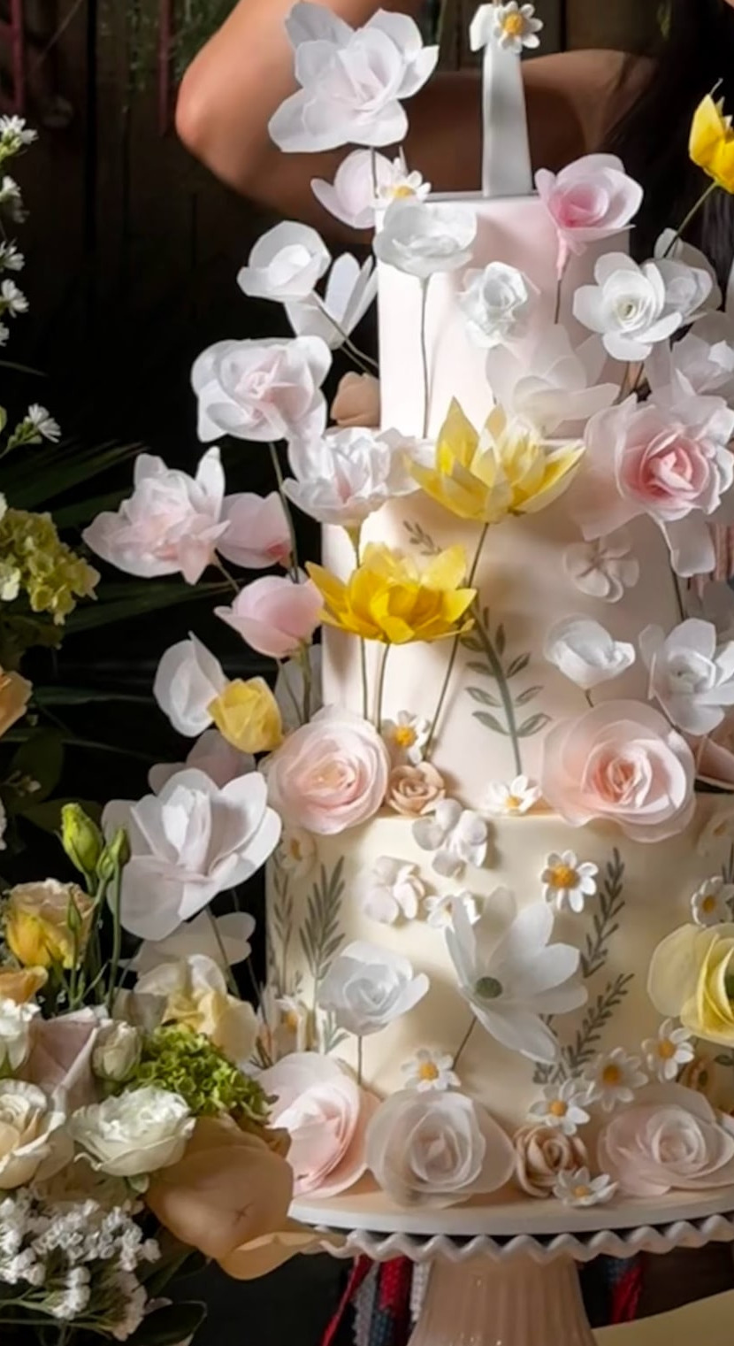 Bolo branco de três andares coberto por rosas, margaridas e tulipas de açúcar em branco, rosa claro e amarelo, com folhagens pintadas à mão