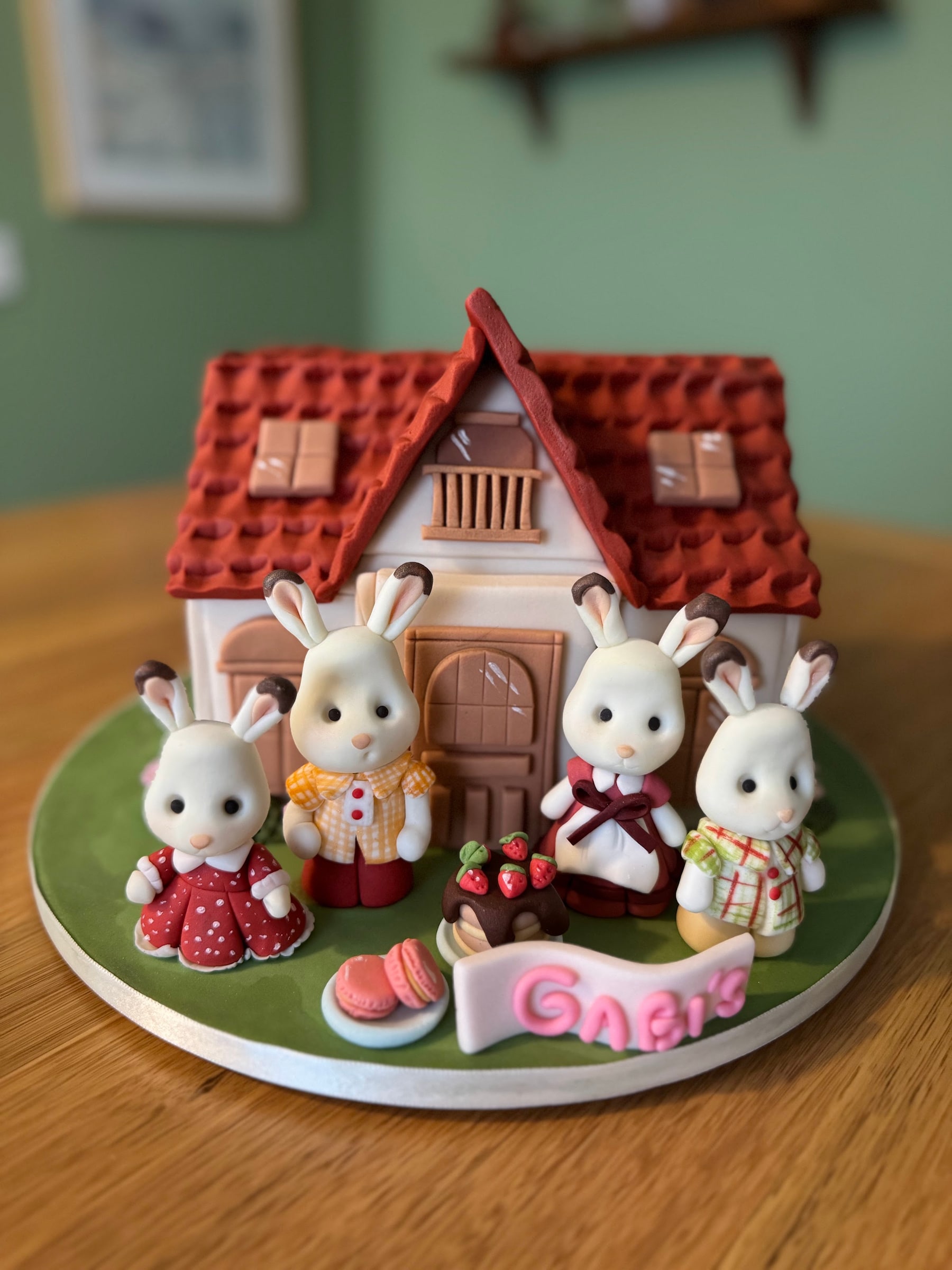 Bolo em formato de casinha de telhado vermelho com quatro coelhos modelados à mão em roupas de época, macarons e um bolinho de morango na base verde