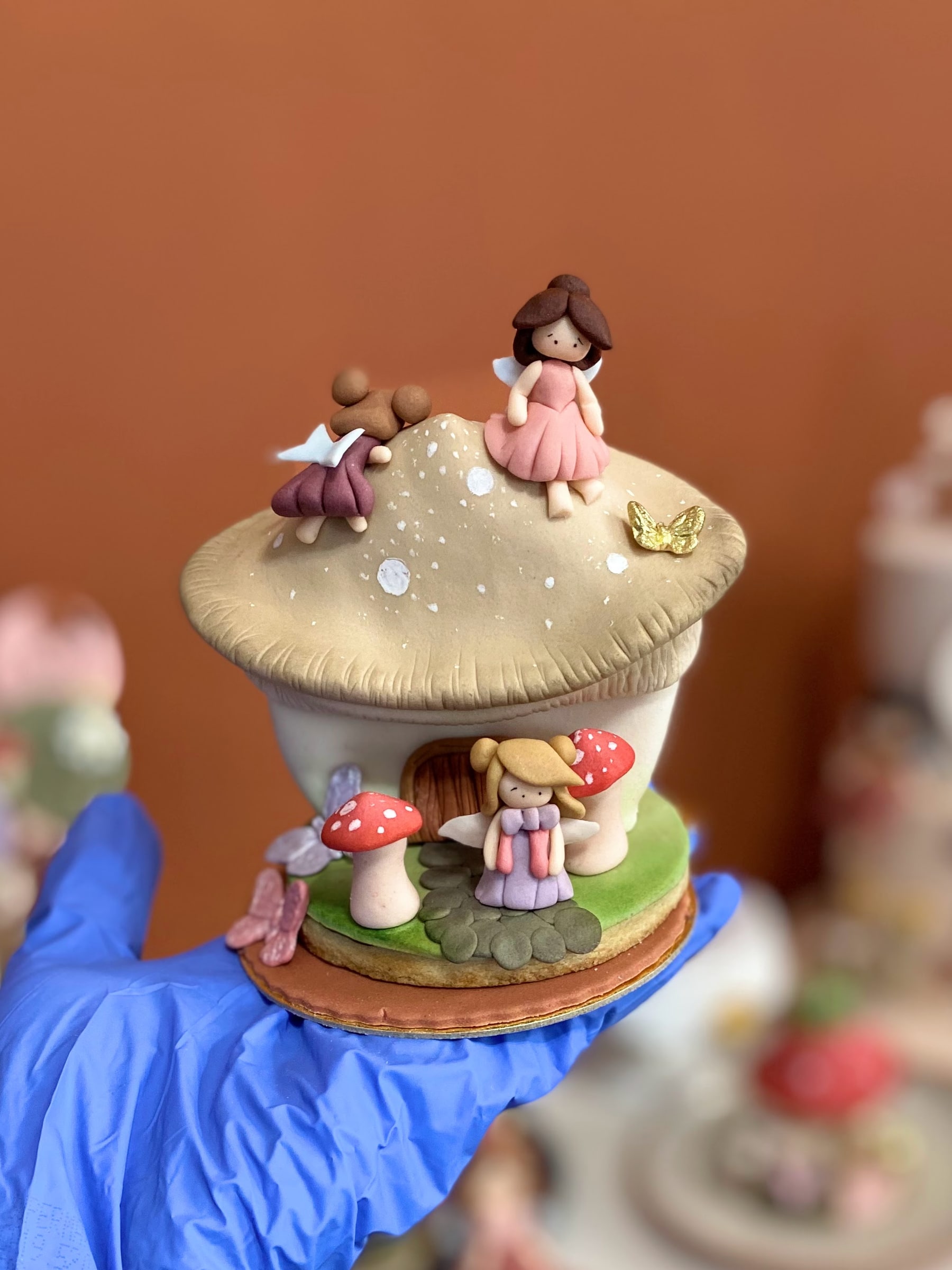 Peça em formato de casa de cogumelo com fadas modeladas à mão, borboleta dourada e cogumelos vermelhos, segurada pela confeiteira