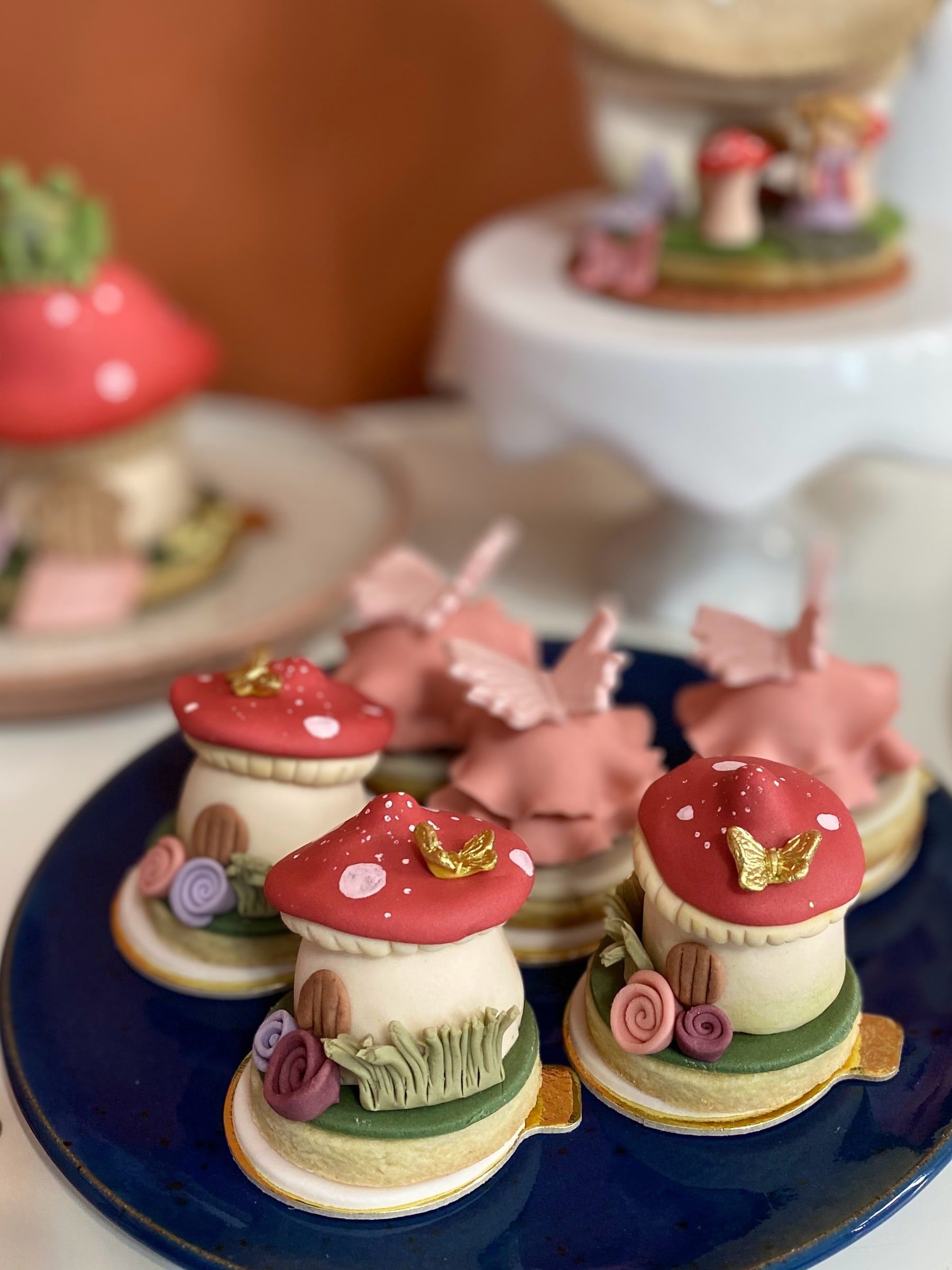 Doces individuais em formato de casa de cogumelo, com chapéu vermelho pintado, borboleta dourada e roseiras modeladas, sobre base de biscoito em prato azul