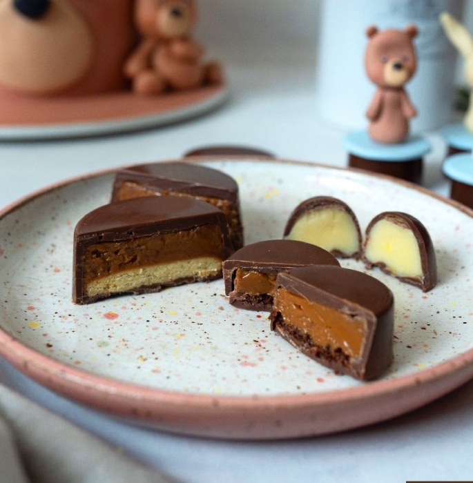 Bombons de chocolate cortados ao meio revelando recheios de doce de leite e creme, em prato de cerâmica rosa salpicado