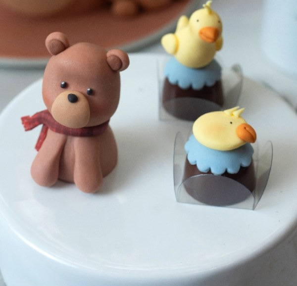 Urso de cachecol e dois patinhos modelados à mão em pasta americana, apoiados sobre bombons de chocolate