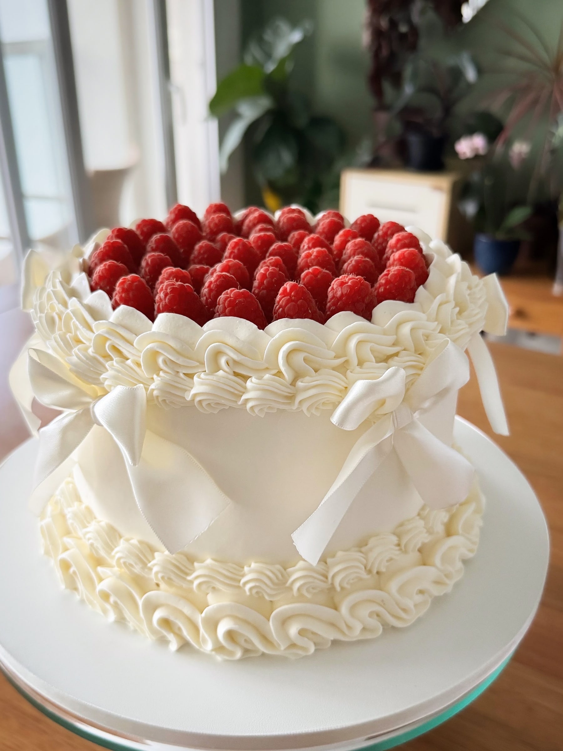 Bolo branco coberto de framboesas frescas no topo, com bordas de buttercream em bicos e laços de cetim marfim ao redor
