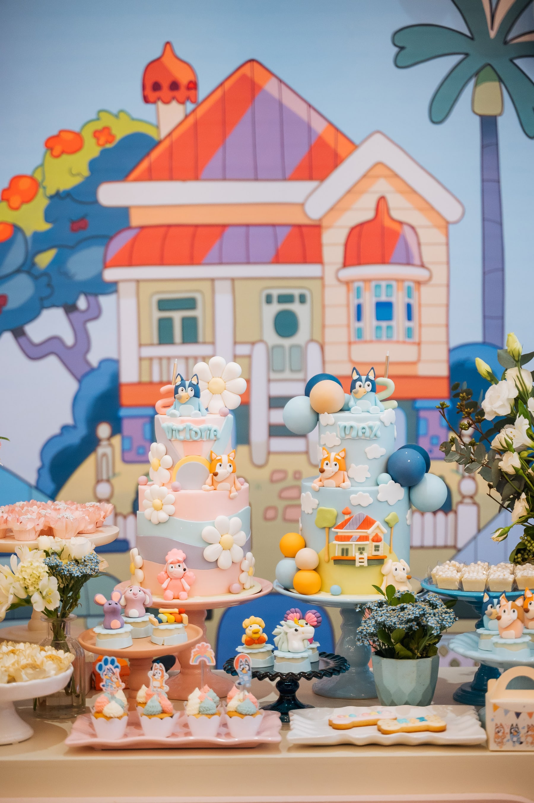 Mesa de festa infantil com dois bolos de dois andares em tons pastel, docinhos modelados, cupcakes e biscoitos decorados, diante de um painel ilustrado