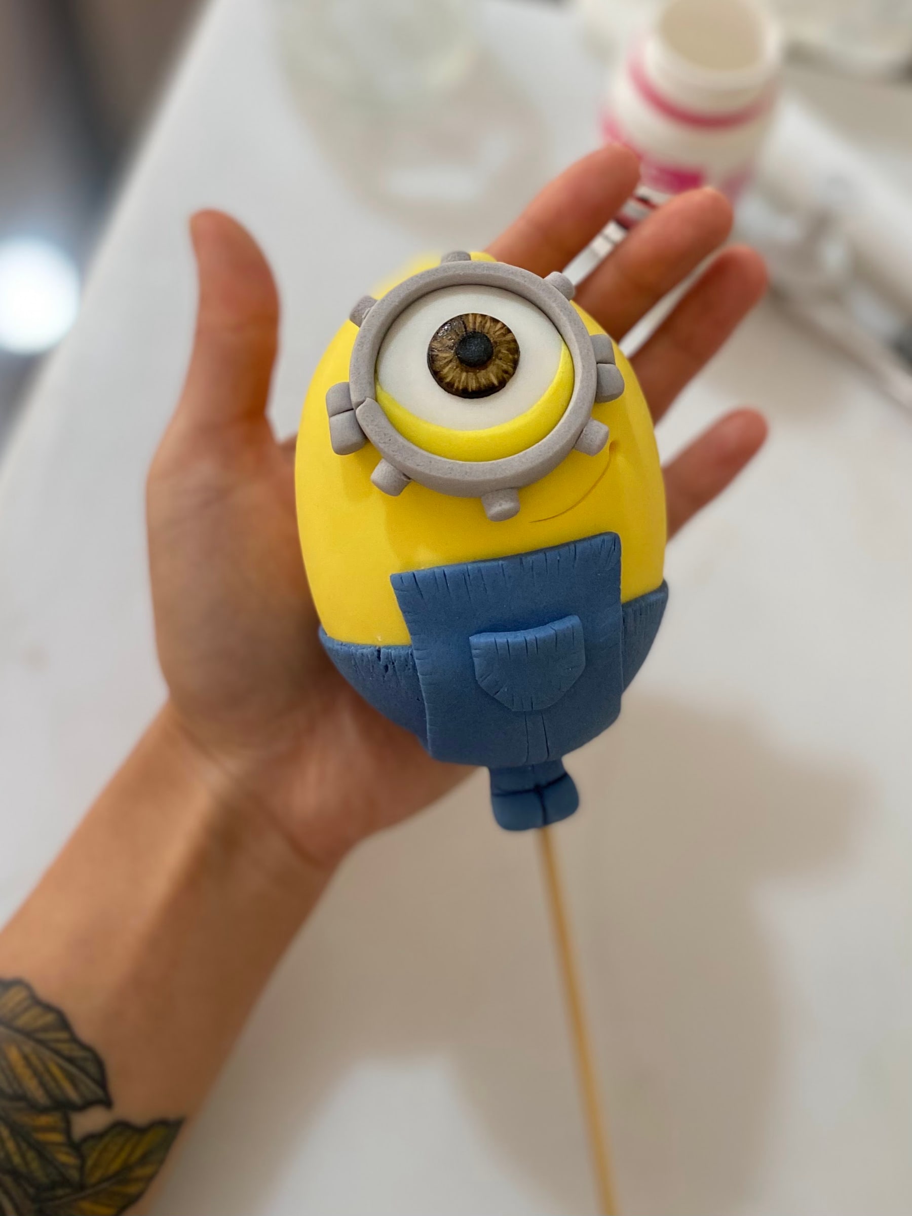 Topo de bolo modelado à mão em pasta americana, ainda sendo segurado pela confeiteira no ateliê, com o olho pintado em detalhe