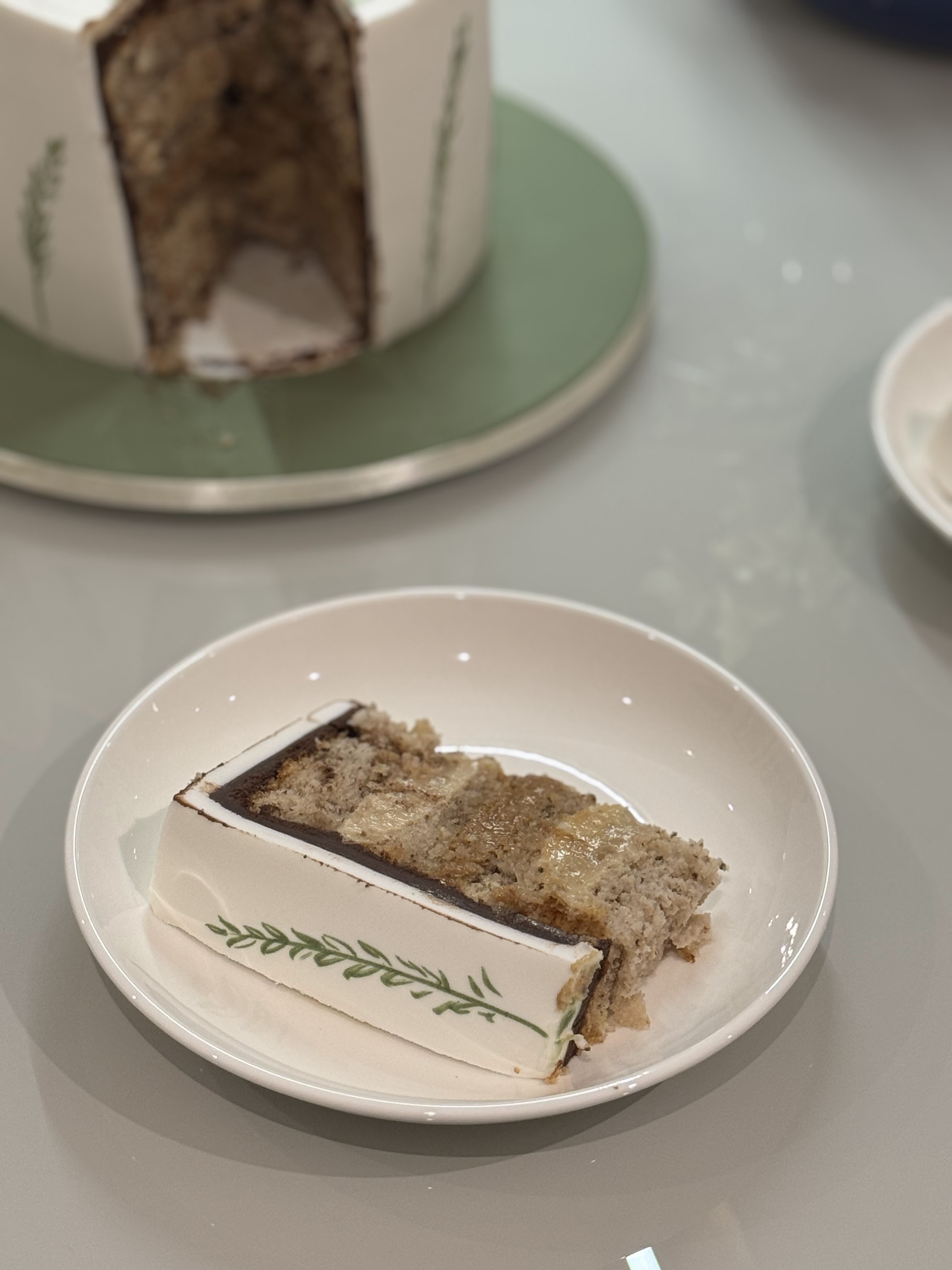 Fatia do bolo de orquídeas em prato de cerâmica, revelando camadas de massa clara, ganache escura e compota dourada, com o ramo botânico pintado na lateral