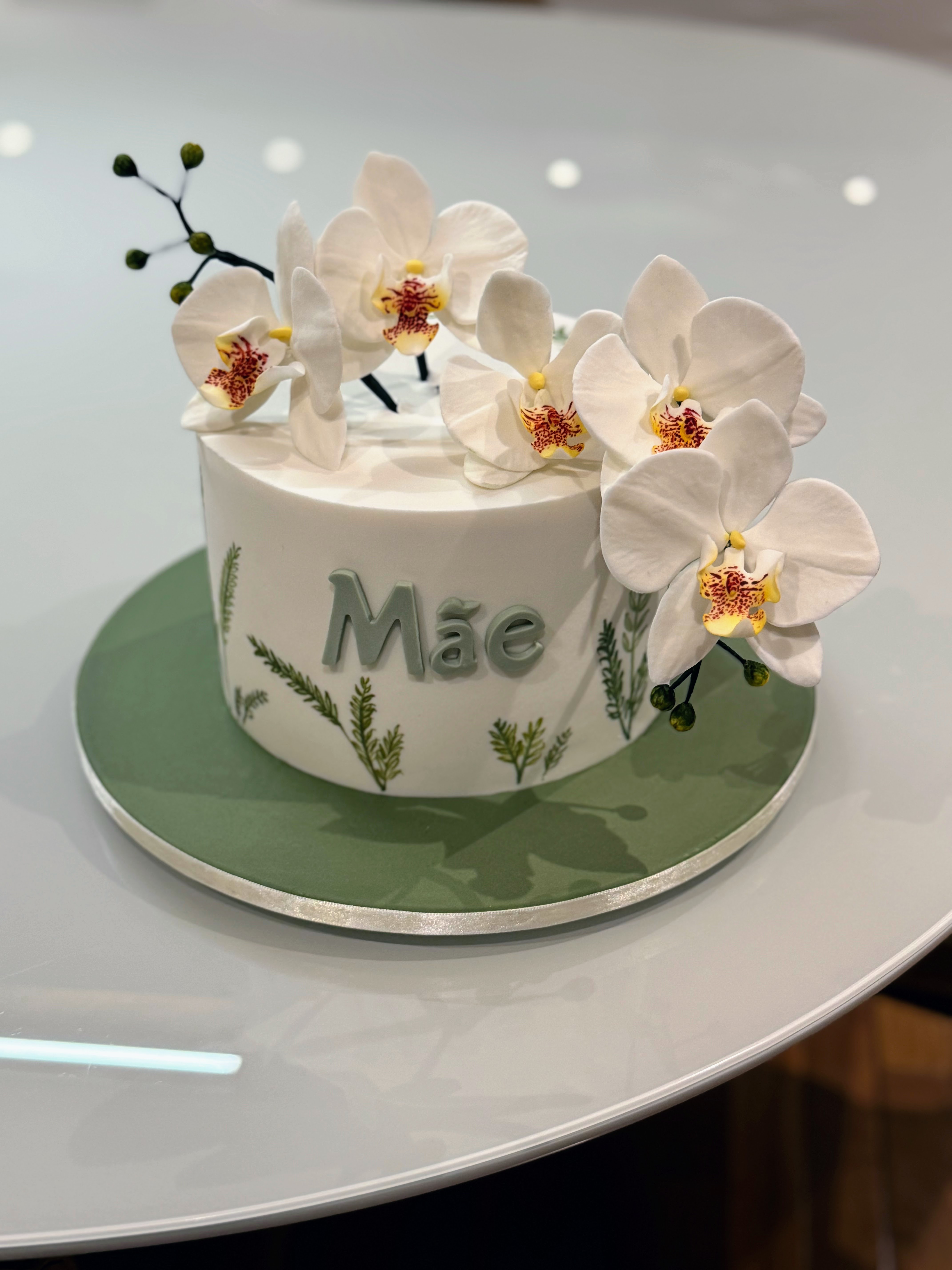 Bolo redondo branco com cinco orquídeas de açúcar brancas abertas no topo, ramos botânicos verdes pintados à mão na lateral e a palavra Mãe em relevo, sobre base verde