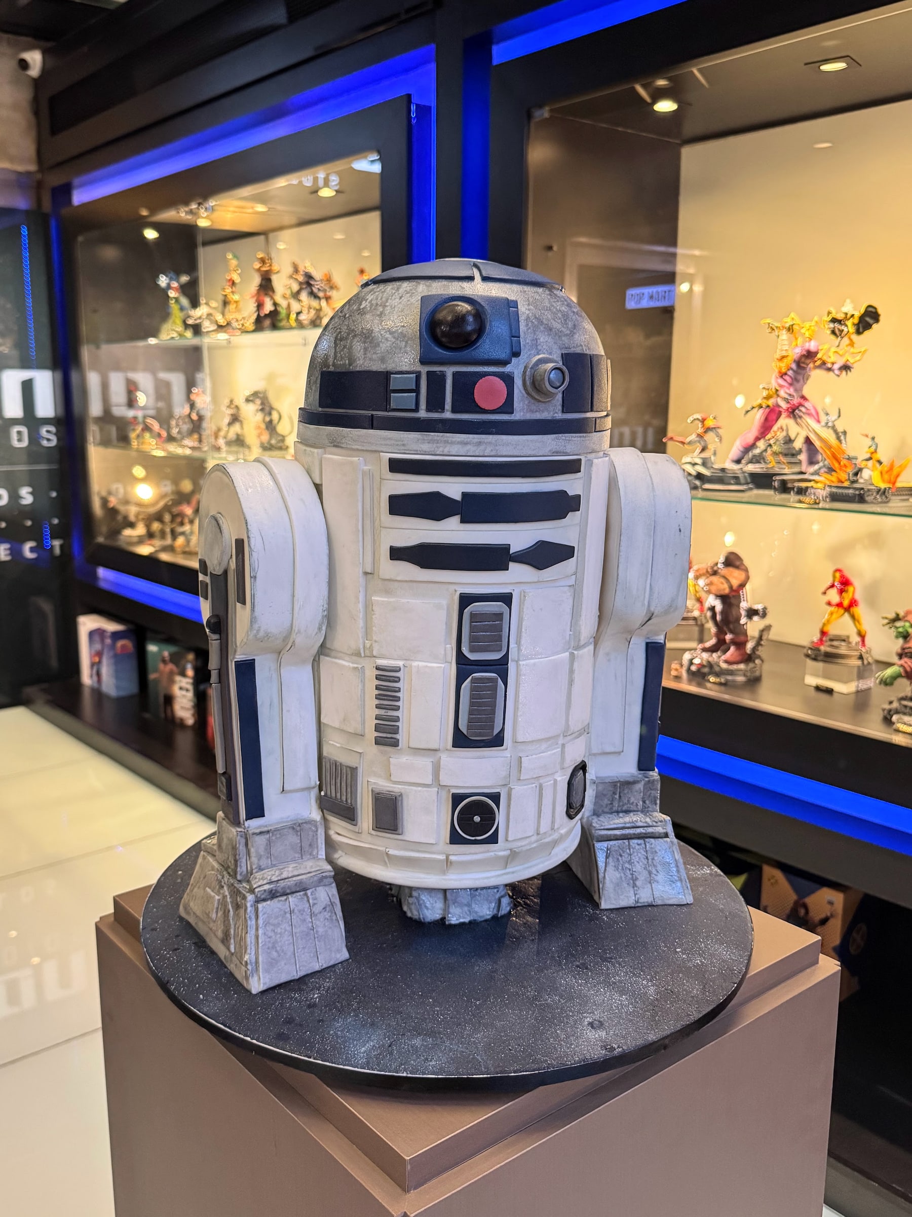 Escultura em bolo do robô R2-D2 em tamanho grande, com acabamento metalizado envelhecido, exposta sobre pedestal em uma loja de colecionáveis