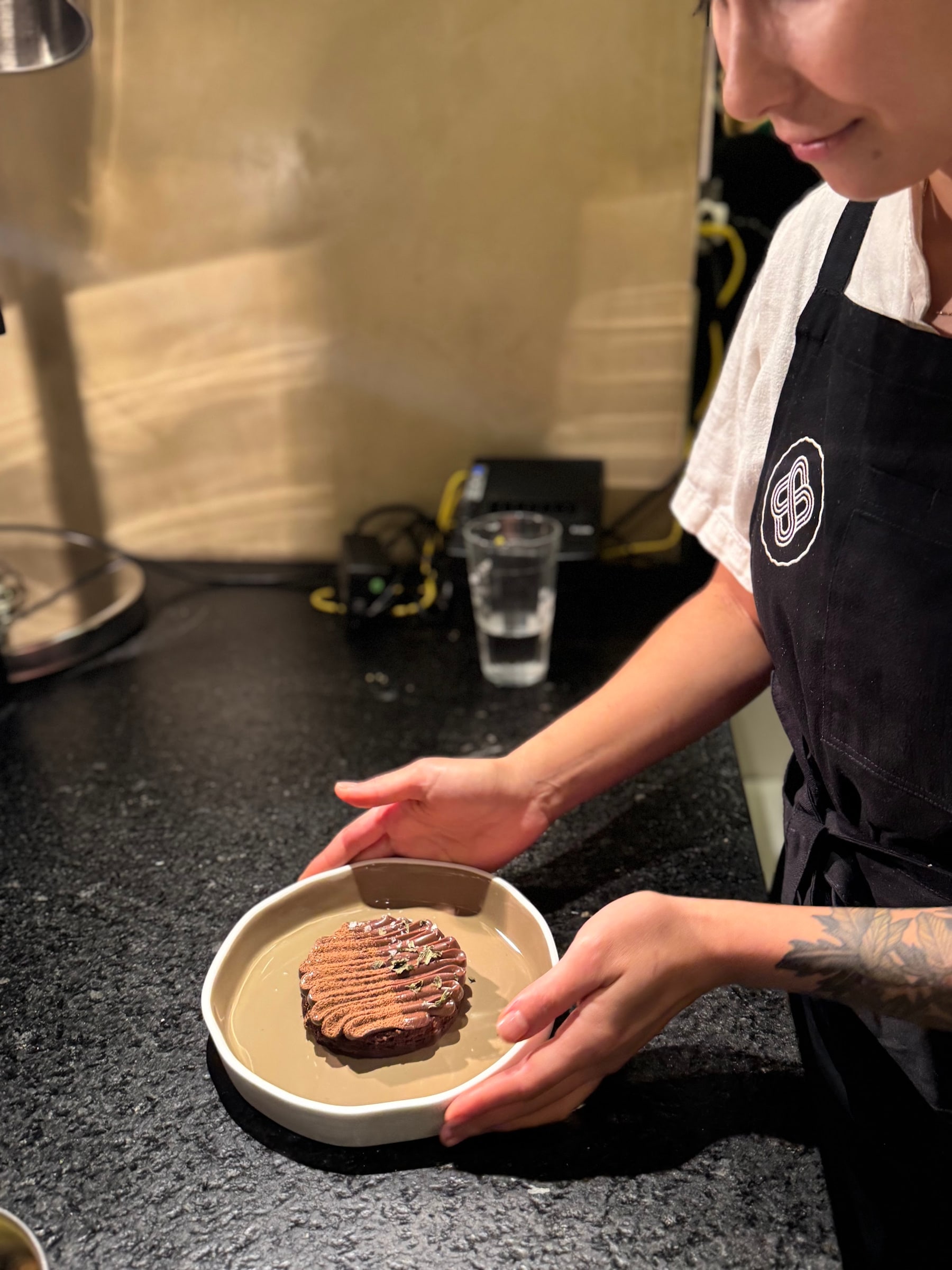Deborah emplatando uma sobremesa de chocolate finalizada em cordões finos, em prato fundo de cerâmica, durante um evento